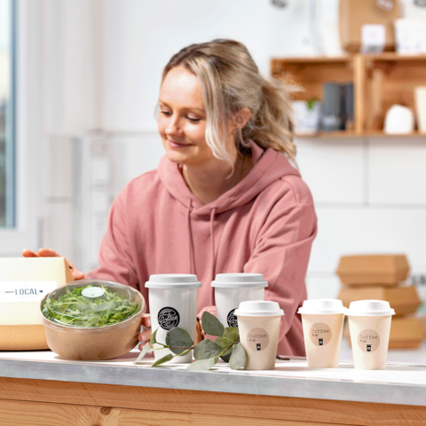 En kvinna i en rosa hoodie förbereder hälsosamma rätter, inklusive en skål med sallad och flera kaffekoppar på ett bord.