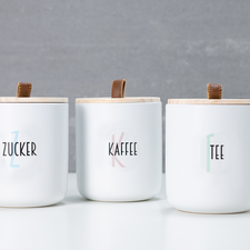 Drei weiße Behälter mit Holzdeckeln, beschriftet mit "Zucker", "Kaffee" und "Tee", auf einem hellen Tisch vor einer grauen Wand.