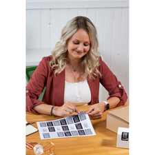 Eine Frau mit blonden, lockigen Haaren sitzt an einem Tisch und betrachtet ein Blatt mit Aufklebern. Neben ihr liegt eine Kiste.