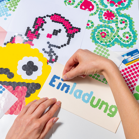 Hände gestalten Einladungen mit bunten Aufklebern auf Papier, darunter ein gelbes Monster und verschiedene Muster.