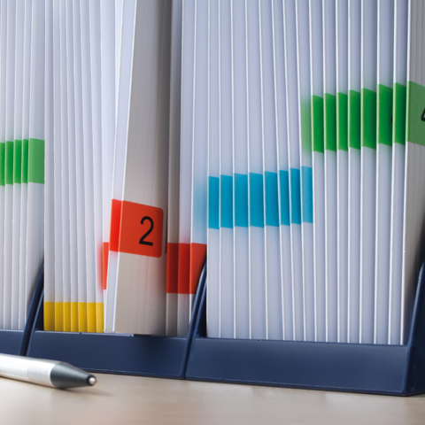 Kartonbox auf einem Tisch mit einem roten Heft und einem grünen "TOP"-Schild. Im Hintergrund sind Bücherregale sichtbar.