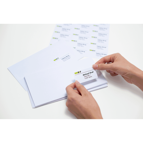 Eine Hand hält ein beschriftetes Kuvert mit dem Namen "Michael Gerd" und einer Adresse, umgeben von leeren Umschlägen und Etiketten.
