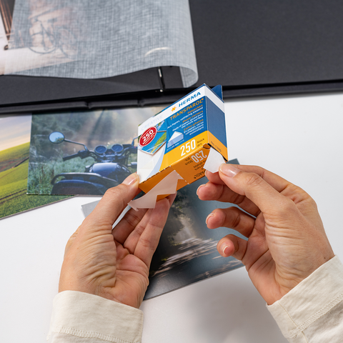 Eine Person hält eine Verpackung mit Fotopapier in den Händen, während mehrere Fotos und ein Album auf einem Tisch liegen.