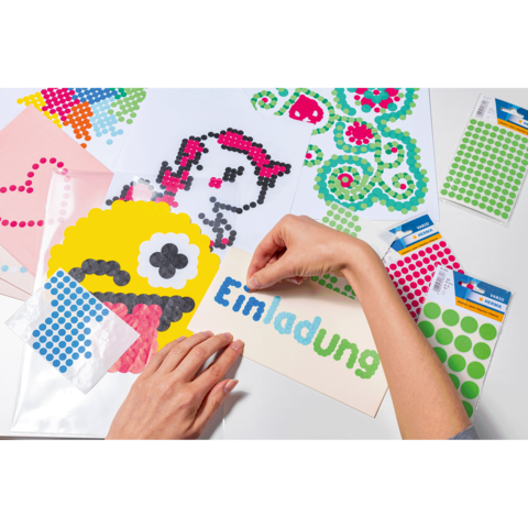 Eine Hand platziert bunte Punkte auf ein Einladungskarten-Design, um ein kreatives Kunstwerk zu gestalten. Verschiedene Aufkleber und Papiere sind sichtbar.
