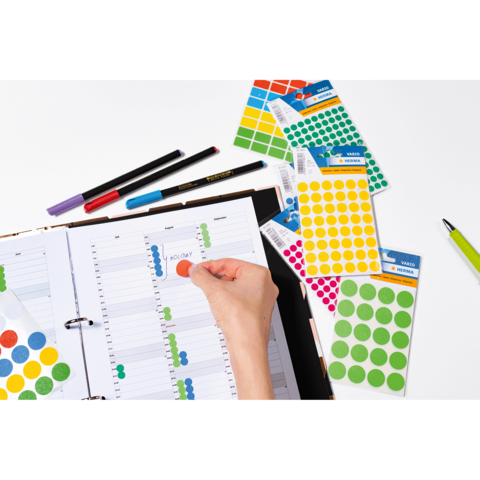 Ein offenes Notizbuch mit einem Kalender, auf dem bunte Aufkleber platziert werden. Stifte und zusätzliche Aufkleberblätter sind sichtbar.