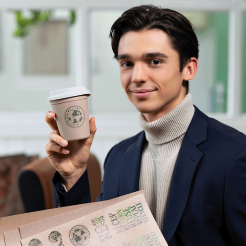 Junger Mann in einem Anzug hält einen Kaffeebecher in der Hand und lächelt, während er vor einem hellen Hintergrund steht.