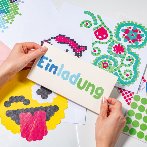 Hände halten eine Einladungskarte mit dem Wort "Einladung" in bunten Buchstaben, umgeben von Bastelmaterialien und Aufklebern.