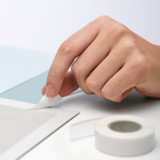 En hand tar bort en bit tejp från en grå yta, medan en rulle tejp ligger bredvid.