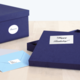 Eine geöffnete, dunkelblaue Geschenkbox mit der Aufschrift "Happy Birthday!" und einem blauen Umschlag daneben. Eine zweite Box steht im Hintergrund.