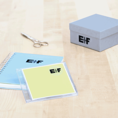 Ein blauer Notizblock, eine gelbe CD-Hülle mit dem Logo "E!F" und eine graue Geschenkbox auf einem Holzuntergrund. Schere daneben.