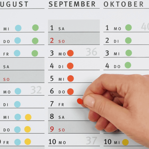 Ein Hand greift nach einem roten Punkt auf einem Kalender, der die Monate August, September und Oktober zeigt, mit hervorgehobenen Daten.