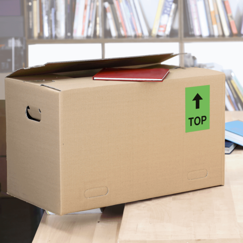 Kartonbox auf einem Tisch mit einem roten Heft und einem grünen "TOP"-Schild. Im Hintergrund sind Bücherregale sichtbar.