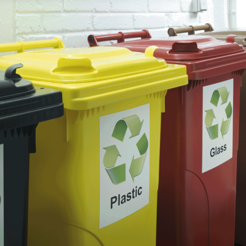 Drei Mülltonnen in verschiedenen Farben mit Recycling-Symbolen: Gelb für Plastik, Rot für Glas und Schwarz für Restmüll.