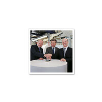 Drei Männer in Anzügen stehen um einen runden Tisch und legen ihre Hände auf ein zentrales Objekt.