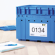 Blaue Kunststoffbox mit der Aufschrift „0134“ und einem Etikett, daneben ein Notizblock und ein blauer Stift auf einem Tisch.