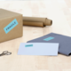 Ein braunes Paket mit dem Aufkleber "FRAGILE", daneben ein blauer Umschlag mit "DRINGEND" und eine Schere auf einem Tisch.