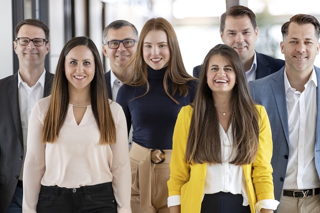 En grupp på sju personer står i en korridor, leende och i formell klädsel, några bär blusar och kostymer.