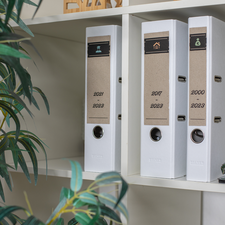 Drei weiße Ordner auf einem Regal, beschriftet mit den Jahren 2020, 2021, 2022 und 2023, neben einem Heftgerät und einer Pflanze.