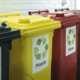 Drei Abfallbehälter in verschiedenen Farben: Gelb für Plastik, Rot für Glas und Schwarz für Restmüll, jeweils mit Recycling-Symbolen.