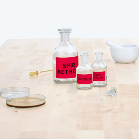 Flasche mit rotem Etikett "Spir. Aether", daneben zwei kleinere Flaschen mit "Barbital" und "Tinct. Tramon", sowie ein Mörser und Petrischalen.