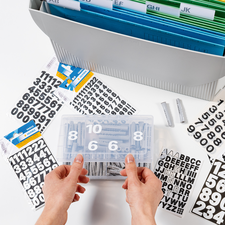 Zwei Hände halten eine transparente Box mit Zahlen und Buchstabenaufklebern, umgeben von weiteren Aufklebern und einem Ordner.
